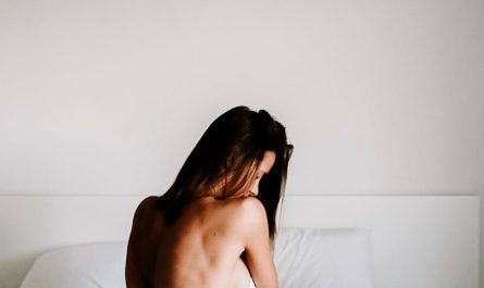 A serene, minimalist portrait of a woman sitting on a bed, enveloped in soft white linens, in a tranquil bedroom setting.